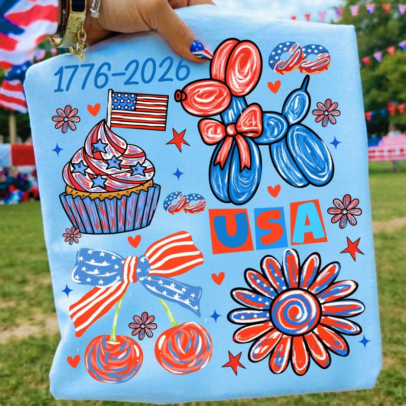 May include: A light blue t-shirt featuring a patriotic graphic print with the text 1776-2026 and USA. The design includes a cupcake with an American flag, a red and blue balloon dog, a pair of cherries with a flag-patterned bow, and a flower.