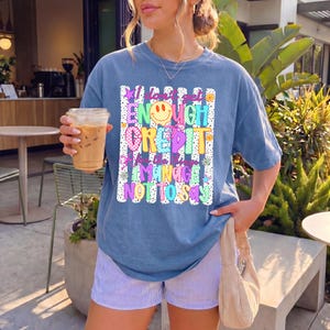 Pode incluir: Uma t-shirt azul de manga curta com um gr&aacute;fico que inclui o texto I don't get ENOUGH CREDIT for the things I MANAGE NOT TO SAY. O design apresenta uma cara sorridente amarela, padr&otilde;es coloridos, estrelas e flores sobre um fundo branco.