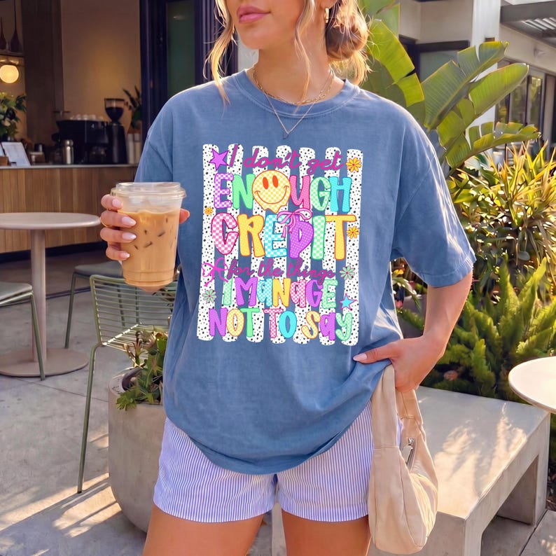 Pode incluir: Uma t-shirt azul de manga curta com um gr&aacute;fico que inclui o texto I don't get ENOUGH CREDIT for the things I MANAGE NOT TO SAY. O design apresenta uma cara sorridente amarela, padr&otilde;es coloridos, estrelas e flores sobre um fundo branco.