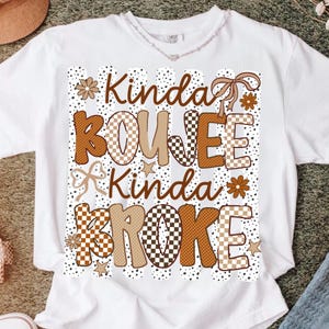 Pode incluir: Uma t-shirt branca de manga curta com uma impress&atilde;o gr&aacute;fica de riscas verticais pretas &agrave;s pintas. O texto Kinda BOUJEE Kinda BROKE apresenta padr&otilde;es de xadrez e bolinhas em castanho e bege, acentuado com pequenas flores, estrelas e la&ccedil;os.