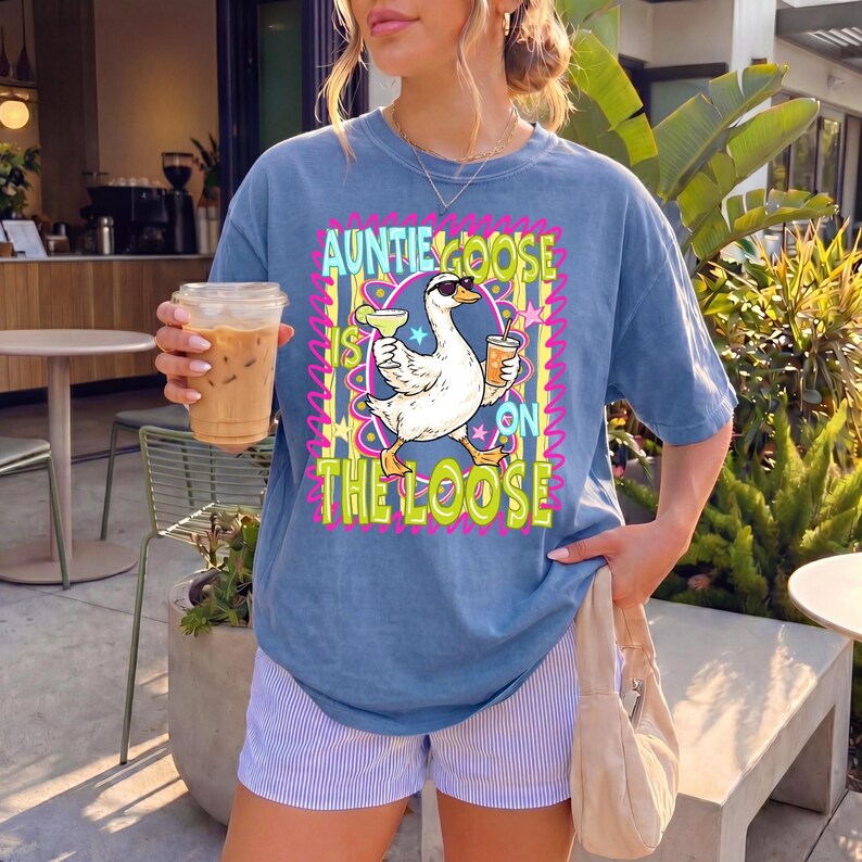 K&ouml;nnte beinhalten: Blaues kurz&auml;rmeliges T-Shirt mit dem Text "Auntie Goose is on the Loose" und einer Cartoon-Gans mit Sonnenbrille, die Cocktails h&auml;lt. Das Shirt wird mit gestreiften Shorts kombiniert. Die Person h&auml;lt ein Getr&auml;nk in einem durchsichtigen Becher.