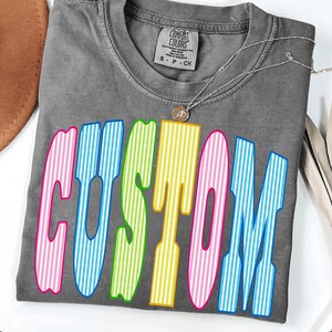 May include: A charcoal gray short-sleeve t-shirt with a crew neckline and a Comfort Colors brand label. The front features the word CUSTOM in large, bold, vertical-striped letters in alternating colors of pink, blue, lime green, and yellow.