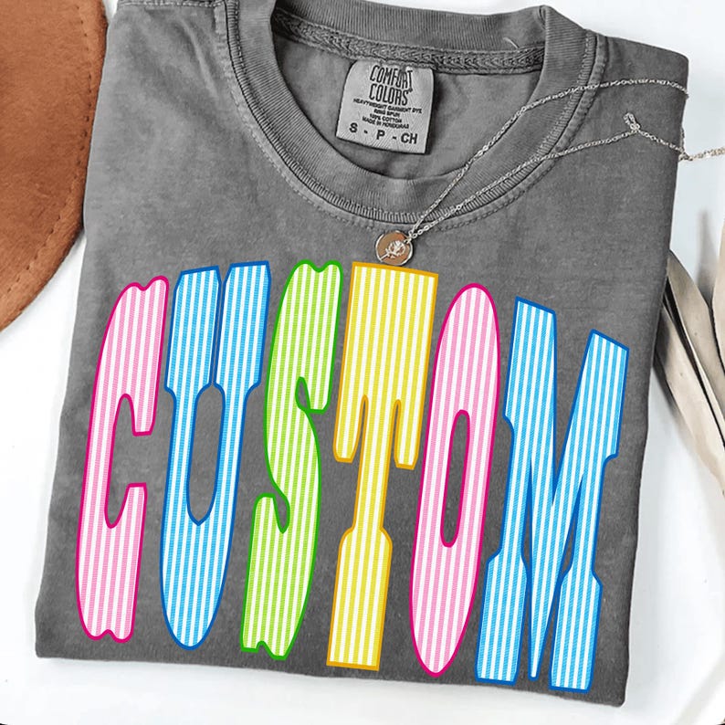 May include: A charcoal gray short-sleeve t-shirt with a crew neckline and a Comfort Colors brand label. The front features the word CUSTOM in large, bold, vertical-striped letters in alternating colors of pink, blue, lime green, and yellow.