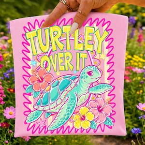 K&ouml;nnte beinhalten: Ein hellrosa Stoffartikel mit einer Grafik einer t&uuml;rkisfarbenen Meeresschildkr&ouml;te, umgeben von rosafarbenen und gelben Hibiskusbl&uuml;ten. Der Text TURTLEY OVER IT ist in gro&szlig;en gelben Buchstaben mit dunkler Umrandung &uuml;ber der Schildkr&ouml;te gedruckt.