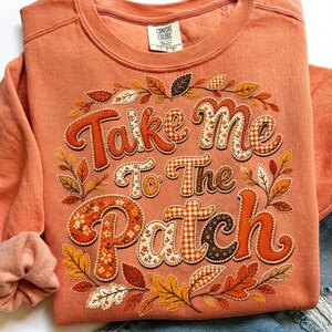 May include: A terracotta-colored crewneck sweatshirt featuring the text "Take Me To The Patch" in a bold, decorative font. The letters are filled with various autumn-themed patterns, surrounded by an embroidered wreath of colorful fall leaves.