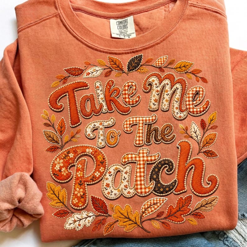 May include: A terracotta-colored crewneck sweatshirt featuring the text "Take Me To The Patch" in a bold, decorative font. The letters are filled with various autumn-themed patterns, surrounded by an embroidered wreath of colorful fall leaves.