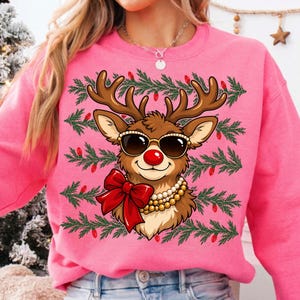 Puede incluir: Una sudadera de color rosa brillante con cuello redondo que presenta un gr&aacute;fico de un reno marr&oacute;n con gafas de sol negras, un lazo rojo grande y un collar de perlas de varias hebras. El fondo incluye filas horizontales de ramas de pino.