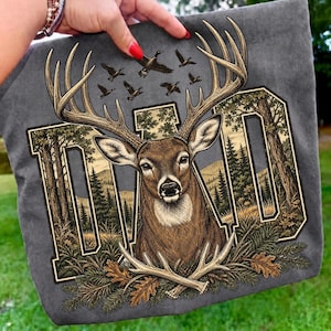 May include: A charcoal grey t-shirt featuring a large graphic of a deer head with prominent antlers. The word DAD is integrated into the background design, which includes a forest landscape, pine trees, and flying ducks. Oak leaves and antlers are at the base.