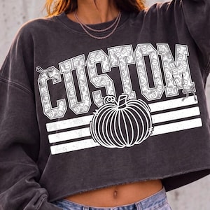 May include: A charcoal grey cropped sweatshirt featuring the word CUSTOM in white block letters filled with a small pumpkin pattern. A large white pumpkin graphic sits below the text, accented by three horizontal white stripes across the chest.