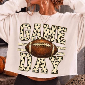 Op de afbeelding: Een cr&egrave;mekleurig sweatshirt met lange mouwen en een grafische print met de tekst GAME DAY in grote blokletters gevuld met zwarte stippen. Een bruine American football met witte veters is tussen de twee woorden in geplaatst.