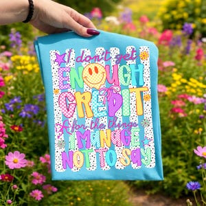 Pode incluir: Uma t-shirt azul brilhante com uma estampa gr&aacute;fica de listras verticais pretas e brancas, um rosto sorridente amarelo e texto colorido que diz: I don't get ENOUGH CREDIT for the things I MANAGE NOT TO SAY, com pequenas estrelas e flores.