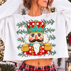 Puede incluir: Una sudadera corta blanca con un estampado gr&aacute;fico de un cascanueces con uniforme rojo y azul verdoso con charreteras doradas. El cascanueces tiene bigote y barba blancos, rodeado de ramas de pino verdes y peque&ntilde;as luces rojas.