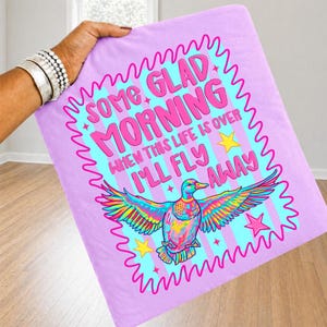 Pu&ograve; includere: Un quadrato di tessuto viola chiaro con una grafica colorata di un'anatra con le ali spiegate. Il testo recita SOME GLAD MORNING WHEN THIS LIFE IS OVER I'LL FLY AWAY. Il design include stelle gialle e un bordo rosa ondulato su sfondo a righe.