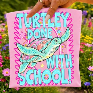 K&ouml;nnte beinhalten: Ein hellrosa Stoffartikel mit einer zentralen Illustration einer Meeresschildkr&ouml;te mit buntem Panzer. Der Text lautet TURTLEY DONE WITH SCHOOL! in gro&szlig;en hellblauen Buchstaben. Das Design enth&auml;lt einen rosa Wellenrand und gelbe Sternakzente.