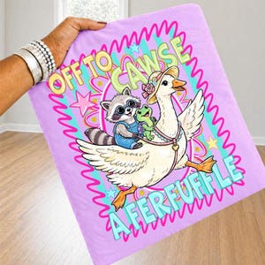 May include: A lavender blanket featuring a whimsical illustration of a goose wearing a hat, carrying a raccoon and a frog. The blanket has the text "OFF TO CAWSE A FERFUFFLE" in a colorful, playful font.