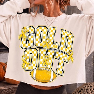 Puede incluir: Una sudadera de manga larga color crema con el texto GOLD OUT en letras grandes de bloque con un patr&oacute;n de tablero de ajedrez amarillo y blanco. El dise&ntilde;o incluye cuatro lazos amarillos de concienciaci&oacute;n y un gr&aacute;fico de f&uacute;tbol americano.