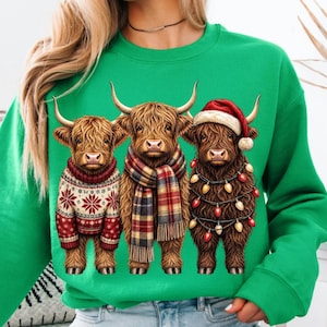 May include: A bright green sweatshirt featuring a graphic of three brown Highland cows. The left cow wears a red and white snowflake sweater, the middle cow wears a plaid scarf, and the right cow wears a Santa hat and string of Christmas lights.
