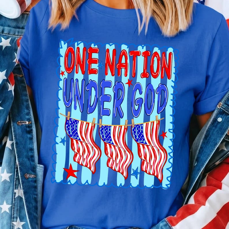 May include: A royal blue t-shirt featuring a graphic print with the text ONE NATION UNDER GOD in red and blue letters. The design includes three American flags hanging from a clothesline against a background of vertical light blue stripes.