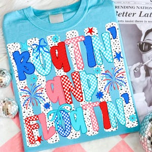 Op de afbeelding: Een lichtblauw t-shirt met de tekst "BOATIN' AND FLOATIN'" in een veelkleurig, gedessineerd lettertype. Het ontwerp bevat rode, blauwe en roze letters met stippen- en dambordpatronen, geaccentueerd door kleine sterretjes, vuurwerk en strikjes.