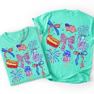 May include: A mint green t-shirt featuring a repeating patriotic graphic print in red, white, and blue. The design includes the Statue of Liberty, American flags, hot dogs, popsicles, fireworks, and bows, all rendered in a stylized, colorful illustration.