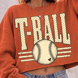 Pode incluir: Uma camisola curta de cor laranja queimado com o texto T-BALL em letras grandes de cor creme com um padr&atilde;o de pontos pretos. Abaixo do texto, h&aacute; um gr&aacute;fico de uma bola de basebol cor creme com costuras pretas e um pequeno cora&ccedil;&atilde;o preto.