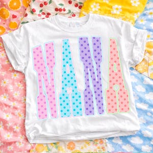 May include: White t-shirt with the word "NANA" in large, colorful, polka-dotted letters. The letters are outlined in light blue and green, with pink, blue, and purple polka dots. The shirt is on a patterned background.