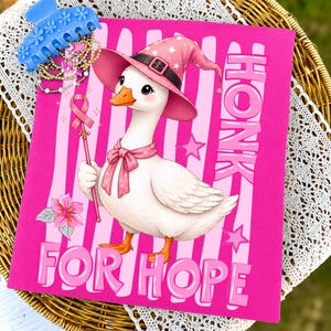 Puede incluir: Una camiseta rosa brillante con una oca blanca que lleva un sombrero de bruja rosa y un lazo rosa. La oca sostiene una varita con un lazo de concienciación rosa. El texto HONK FOR HOPE aparece en letras grandes sobre rayas y estrellas rosas.