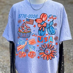 May include: A light grey short-sleeved t-shirt featuring a patriotic graphic with the text 1776-2026 and USA. The design includes a cupcake with an American flag, a red and blue balloon dog, a pair of cherries with a striped bow, and a flower.
