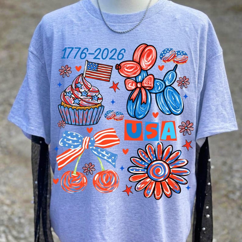 May include: A light grey short-sleeved t-shirt featuring a patriotic graphic with the text 1776-2026 and USA. The design includes a cupcake with an American flag, a red and blue balloon dog, a pair of cherries with a striped bow, and a flower.