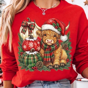 May include: A red crewneck sweatshirt featuring a graphic of a fluffy Highland cow wearing a Santa hat and a plaid scarf, surrounded by three decorative pumpkins, pine branches, pinecones, and red berries in a festive holiday arrangement.