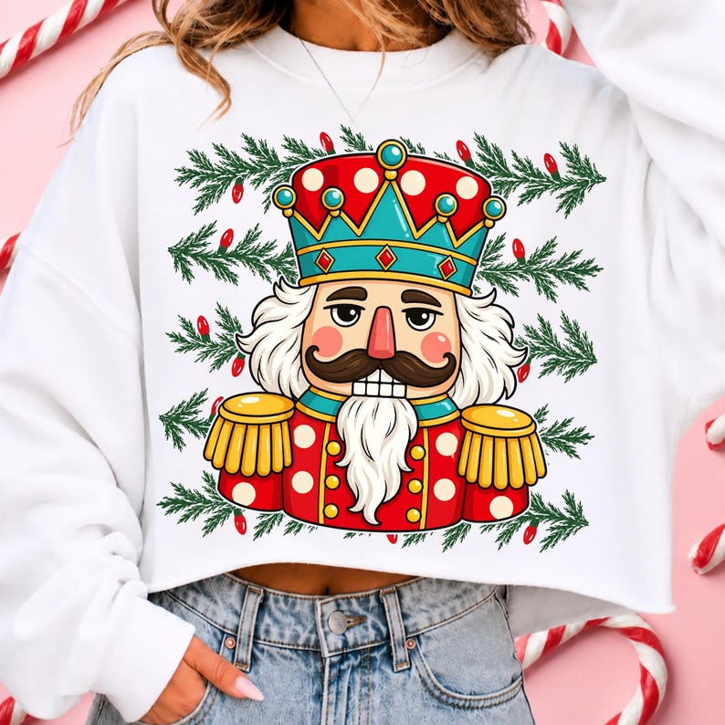 Puede incluir: Una sudadera corta blanca con una ilustraci&oacute;n gr&aacute;fica de un cascanueces que lleva una corona y un uniforme rojo y azul turquesa con charreteras doradas. El dise&ntilde;o incluye un fondo de ramas de pino verdes y peque&ntilde;as luces navide&ntilde;as rojas.