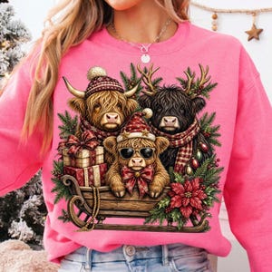 Puede incluir: Una sudadera rosa brillante con un gr&aacute;fico de tres vacas Highland en un trineo de madera. Las vacas llevan gorros de invierno, bufandas y gafas de sol, rodeadas de ramas de pino, flores de pascua rojas, adornos navide&ntilde;os y regalos.