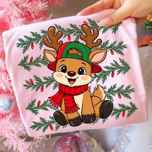 May include: A light pink t-shirt featuring a cartoon reindeer wearing a green and red baseball cap and a red scarf. The reindeer is centered against a background of green pine branches and small red Christmas lights, creating a festive design.