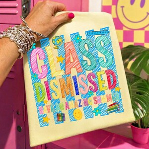 May include: A cream-colored tote bag featuring a colorful graphic with the text "CLASS DISMISSED" and "HAVE AN AMAZING SUMMER." The design includes blue diagonal stripes, a globe, flowers, stars, a smiley face, crayons, and pencils.