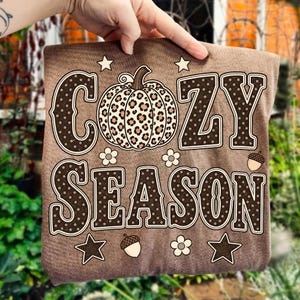 Może przedstawiać: Brązowy t-shirt z napisem COZY SEASON wykonanym ciemnobrązowymi literami w białe kropki. Pośrodku między słowami znajduje się dynia w panterkę. Wz&oacute;r zawiera małe białe kwiatki, gwiazdki oraz żołędzie.