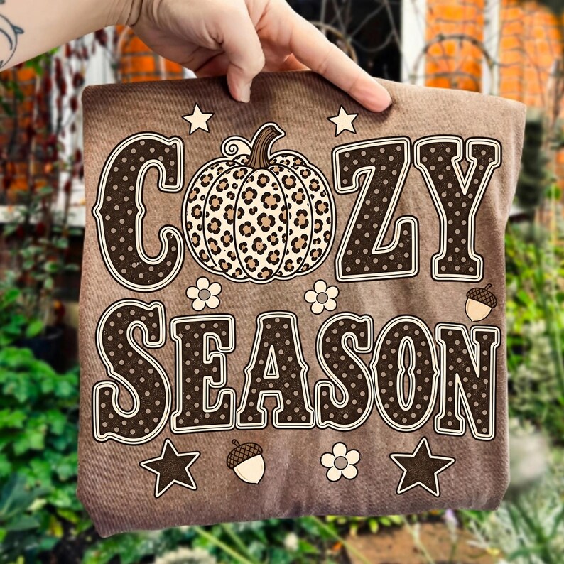 Może przedstawiać: Brązowy t-shirt z napisem COZY SEASON wykonanym ciemnobrązowymi literami w białe kropki. Pośrodku między słowami znajduje się dynia w panterkę. Wz&oacute;r zawiera małe białe kwiatki, gwiazdki oraz żołędzie.