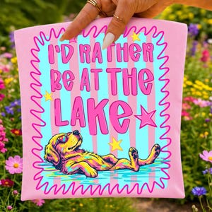 May include: A pink rectangular fabric item featuring a light blue and pink striped background with a pink squiggly border. The text reads I'D RATHER BE AT THE LAKE in pink letters, accompanied by yellow stars and a golden dog wearing sunglasses.