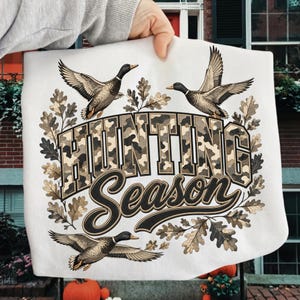 May include: A white sweatshirt featuring a graphic print of three flying mallard ducks surrounded by oak leaves. The text "HUNTING SEASON" is displayed in the center, with "HUNTING" in a camouflage-patterned block font and "Season" in black script.
