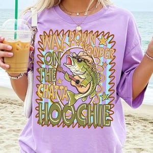 May include: A lilac-colored t-shirt featuring a cartoon bass fish wearing a cowboy hat and sunglasses, playing a guitar. The shirt has the text "WAY DOWN YONDER ON THE CHATTA HOOCHIE" in a decorative font.
