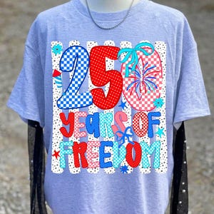 May include: A light gray short-sleeve t-shirt featuring a graphic with the text 250 YEARS OF FREEDOM. The design includes red, white, and blue checkered and polka-dot patterns, firework illustrations, and blue bows against a vertical striped background.