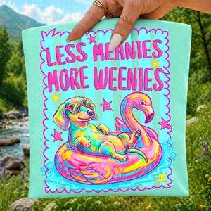 May include: A light mint green tote bag featuring a colorful graphic of a dachshund wearing heart-shaped sunglasses, lounging on a pink flamingo pool float. The text reads LESS MEANIES MORE WEENIES in bold pink letters, surrounded by yellow and pink stars.