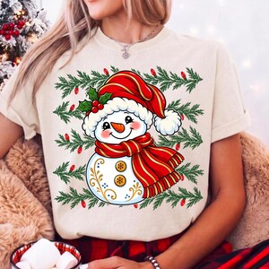 May include: A cream-colored t-shirt featuring a graphic of a smiling snowman wearing a red Santa hat and a red striped scarf. The snowman is decorated with gold swirls and two buttons, set against a background of green pine branches and red lights.