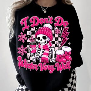May include: A black sweatshirt featuring a graphic of a skeleton wearing a pink knit hat and scarf, holding a pink coffee cup. The design includes pink snowflakes, a leopard print lightning bolt, and the text I Don't Do Winter Very Well.