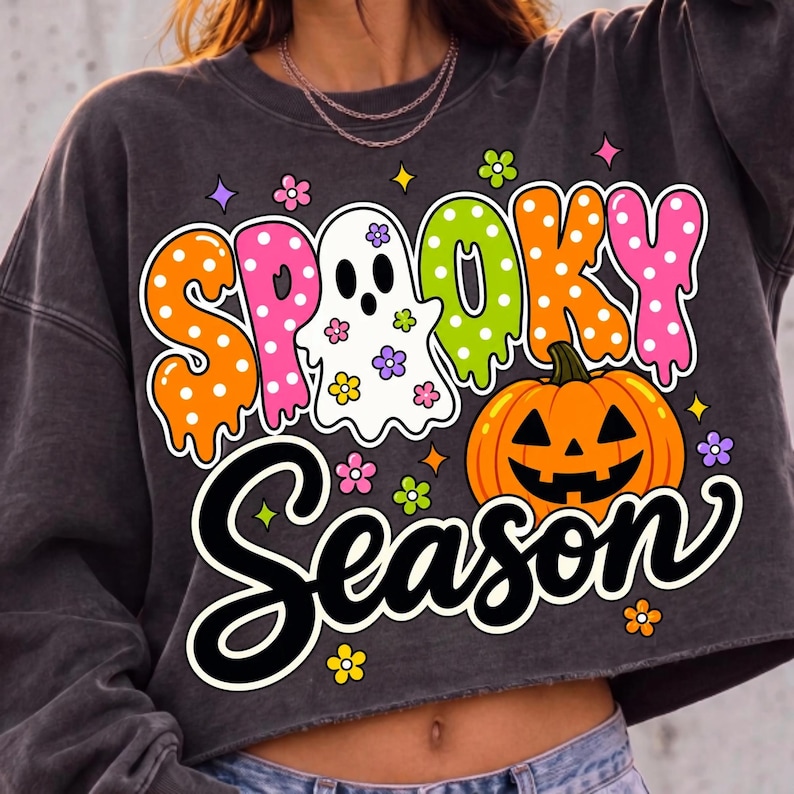 May include: A charcoal gray cropped sweatshirt featuring a graphic print with the text "SPOOKY SEASON". The design includes a white ghost, a bright orange jack-o'-lantern, and colorful polka-dot lettering decorated with small flower icons.