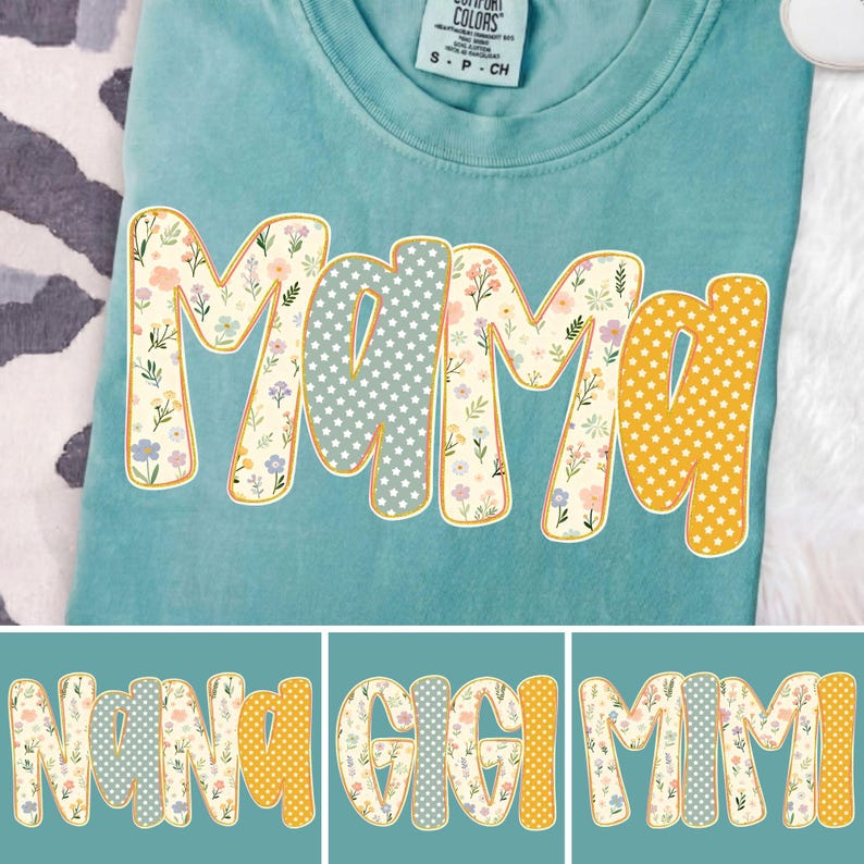 May include: A teal crew neck t-shirt featuring the word MAMA in large, blocky letters. The letters have a cream background with a colorful floral pattern, a light blue background with white stars, and a yellow background with white polka dots.