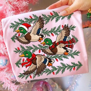 May include: A light pink t-shirt featuring a graphic of three flying mallard ducks. One duck wears a Santa hat, one wears a red and green plaid scarf, and one wears a red bow. The ducks are surrounded by green pine branches and red Christmas lights.