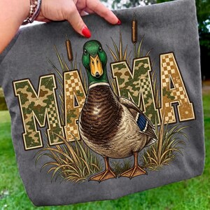 May include: A gray fabric item featuring a graphic of a mallard duck standing among cattails. The word MAMA is printed in large, block-style letters with a camouflage and checkered pattern. The duck has a green head, yellow beak, and brown body.