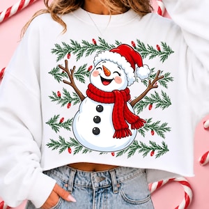 Puede incluir: Una sudadera blanca corta con el gr&aacute;fico de un mu&ntilde;eco de nieve sonriente que lleva un gorro de Pap&aacute; Noel rojo y una bufanda roja. El mu&ntilde;eco de nieve tiene brazos de ramitas y tres botones negros. El dise&ntilde;o incluye ramas de pino verdes y luces rojas.