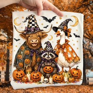 May include: A white t-shirt featuring a graphic illustration of a Highland cow, a raccoon, and a goose wearing witch hats and Halloween capes. The animals are surrounded by pumpkins, bats, crescent moons, stars, and lit candles on the ground.