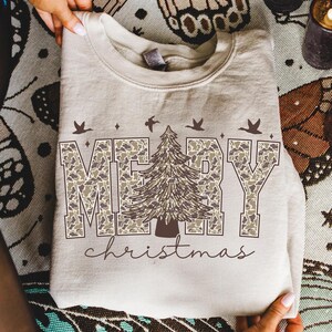May include: A beige sweatshirt with the words "MERRY christmas" printed on the front. The word "MERRY" is in a camouflage pattern, with a detailed Christmas tree in the center. Above the word "MERRY" are three birds.